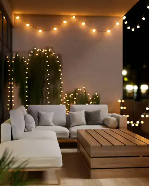 Cozy balcony setup featuring a white sectional sofa with pillows, a wooden coffee table, and string lights creating a warm ambiance.