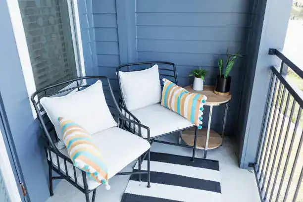 A cozy balcony with two black metal chairs featuring white cushions and colorful striped pillows. A small round table holds a potted plant and a decorative vase, set against a blue wall.