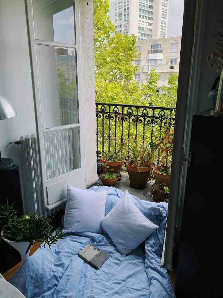 A cozy corner with a blue blanket and pillows on the floor, leading to a balcony with potted plants and a view of tall buildings and green trees.