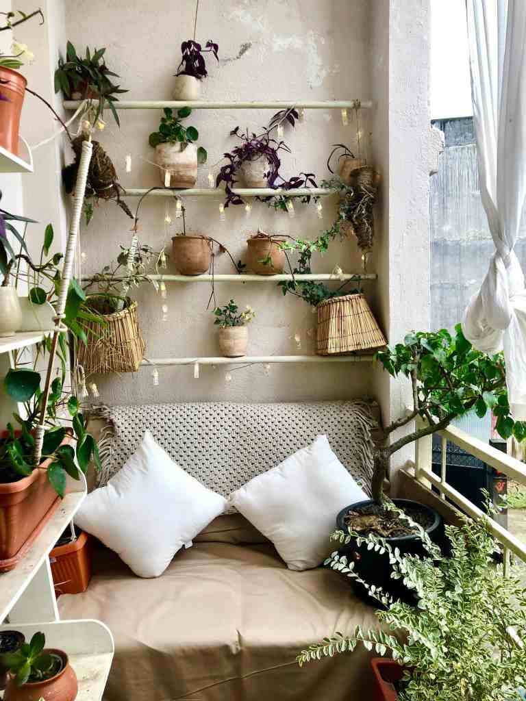 A cozy balcony with a beige wall, featuring shelves adorned with various potted plants and hanging planters. A comfortable seating area with two white pillows on a tan blanket completes the inviting space.