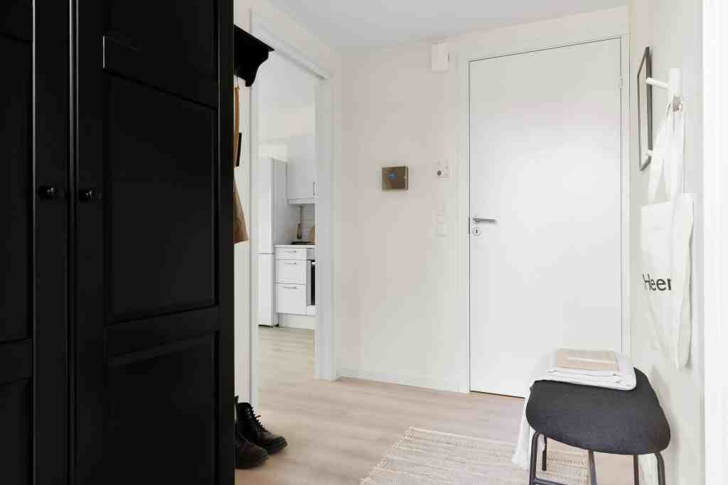 A modern hallway featuring a black wardrobe, a white door, and a small black bench with a folded towel on top. The space has light wooden flooring and leads to a kitchen visible in the background.