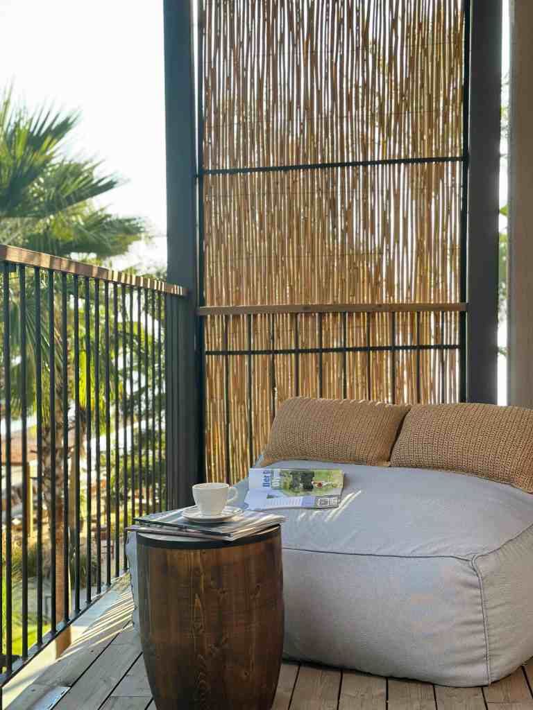 A cozy balcony setup featuring a large bean bag chair with a woven backrest, a wooden side table with a cup and magazine, and bamboo fencing, surrounded by palm trees.