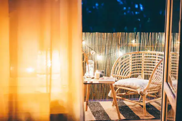 Cozy outdoor balcony setting at night featuring a wicker chair, a small table with candles, and soft lighting in the background.