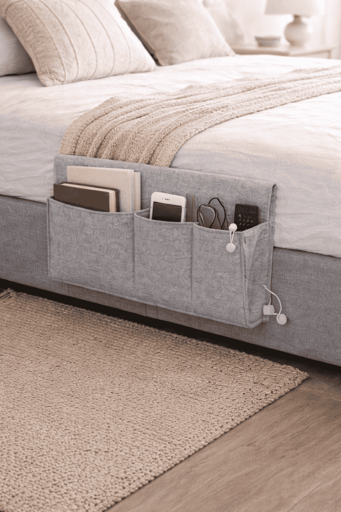 A bedside storage organizer hung on a bed, featuring multiple pockets that hold books, a smartphone, a remote control, and earbuds, with a cozy bedroom setting in the background.