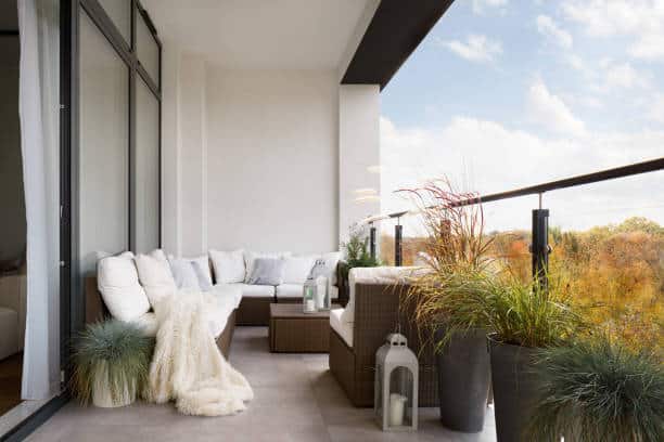 A modern balcony with stylish seating, cozy throw blankets, and potted plants, featuring a view of trees in autumn.