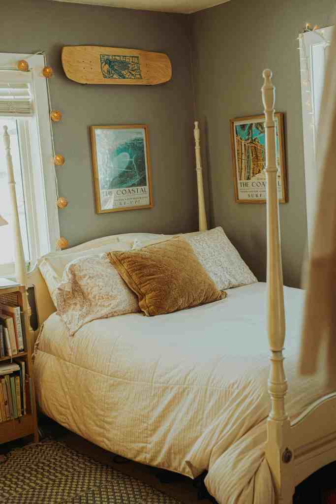 Cozy bedroom featuring a white bed with decorative pillows, a wooden skateboard on the wall, and framed coastal-themed art. A bookshelf is visible beside the bed, and soft lighting adds warmth to the room.
