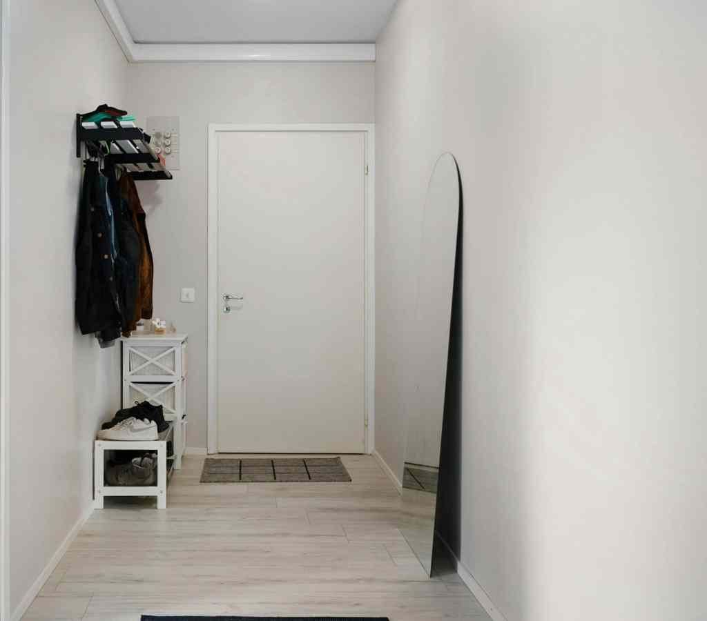 A modern hallway featuring a white wall, a closed door, a full-length mirror, and a small storage unit with shoes and a rug on the floor.
