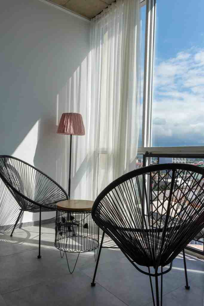 A cozy corner with two black chairs, a wooden table, and a pink lamp, illuminated by natural light from large windows, showcasing a city view.