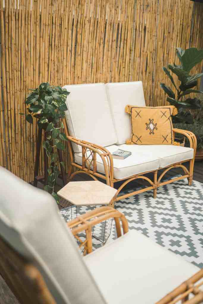 A cozy seating area featuring a rattan sofa with white cushions and a decorative yellow pillow, surrounded by potted plants and a bamboo wall, with a geometric patterned rug and a small hexagonal table.