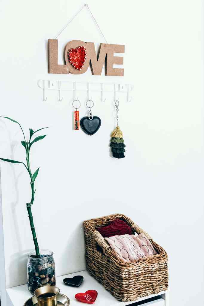 A decorative wall with a 'LOVE' sign featuring a red heart, multiple keychains hanging from hooks, a bamboo plant in a jar, and a woven basket filled with colorful knitted items.