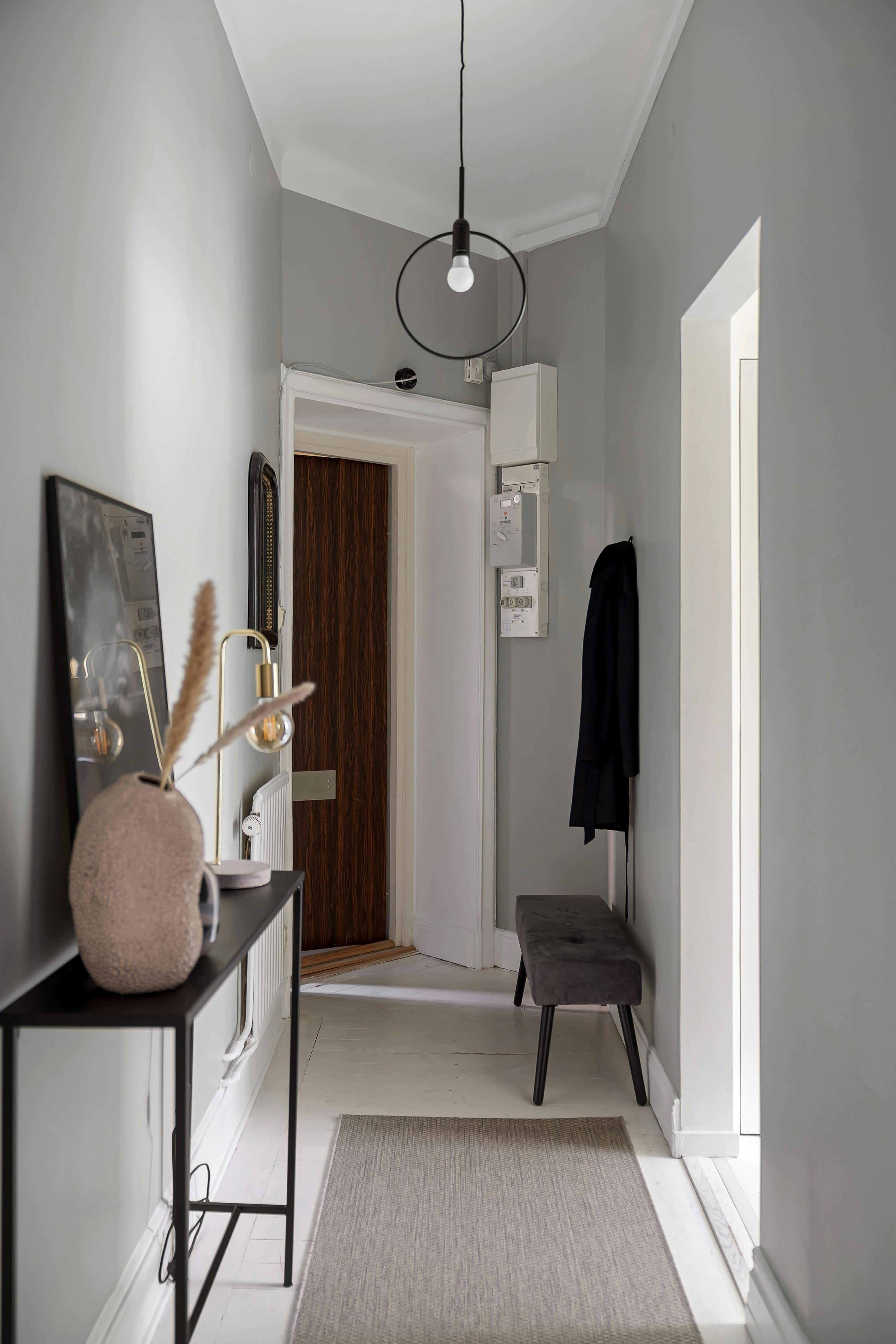 A modern hallway featuring gray walls, a pendant light, a console table with decorative items, a dark wooden door, and a bench.