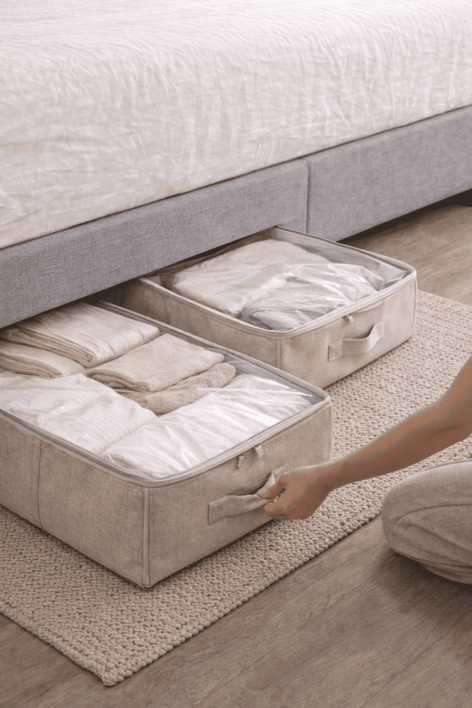 A person organizing clothing in storage bins under a bed, with neatly folded items visible inside the bins.